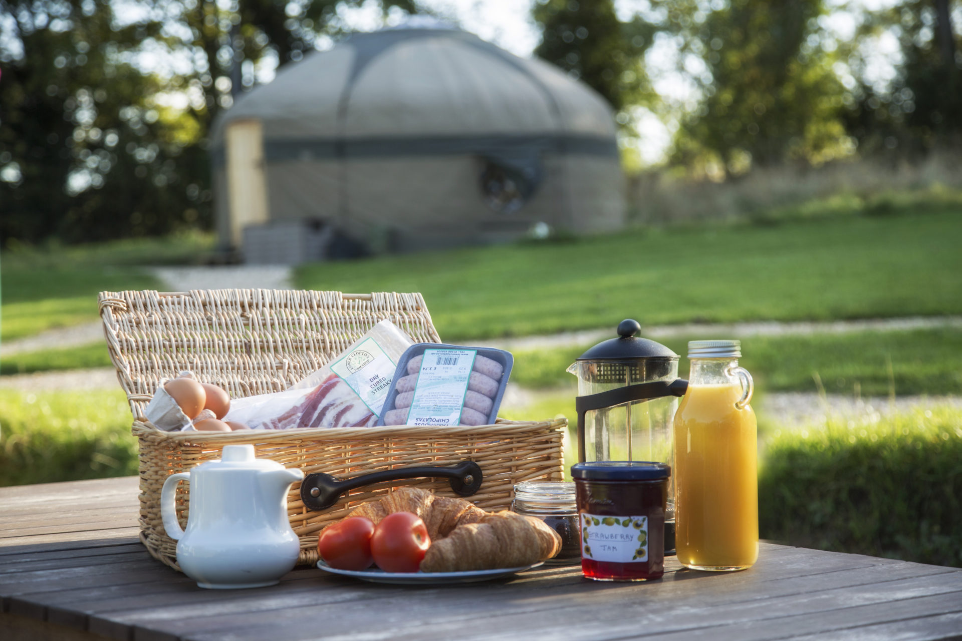 Breakfast hamper with sausages, bacon, eggs and drinks on table in front of yurt