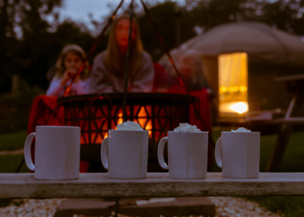 4 mugs of hot chocolate in the foreground with campfire and yurt in background